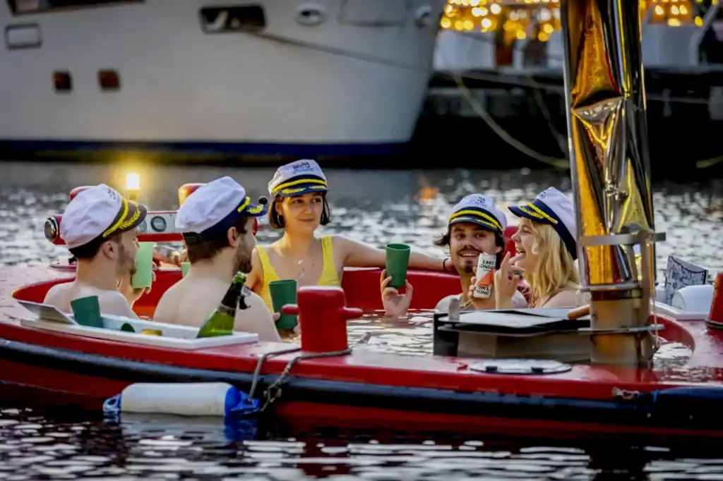 People relaxing in a floating hot tub with green cups while drifting along the River Thames near Canary Wharf on a sightseeing boat.