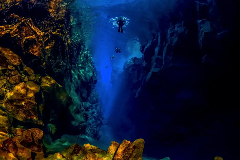 Deep Silfra Fissure in Iceland shows crystal-clear blue-green water and rocky walls, with divers drifting in the gap between tectonic plates.