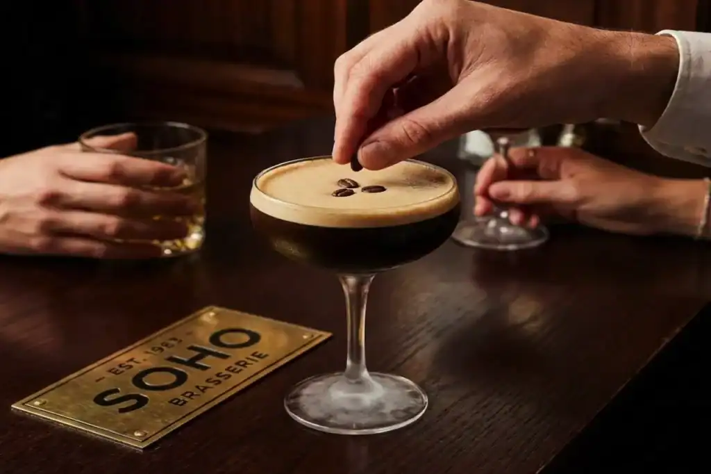 A bartender’s hand lowers three coffee beans onto the foamy crema of an espresso martini, continuing the notorious “wake me up” ritual.