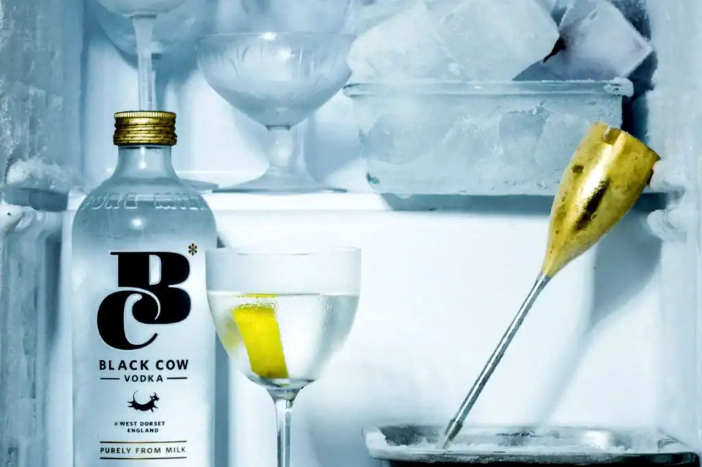 Chilled Black Cow Vodka milk vodka from West Dorset sits beside a glass cocktail, with a gold-tipped pour and cool blue ice backdrop.