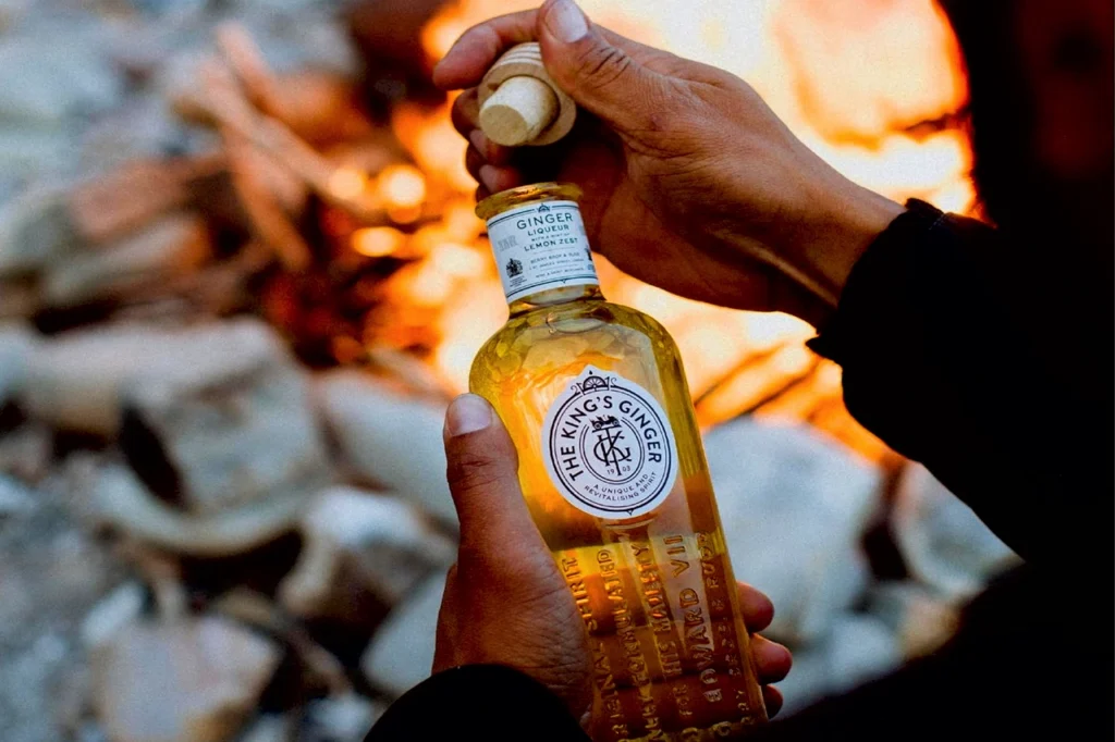 A hand holds an amber glass bottle labeled King’s Ginger ginger liqueur, reflecting Berry Bros. & Rudd’s royal formula.