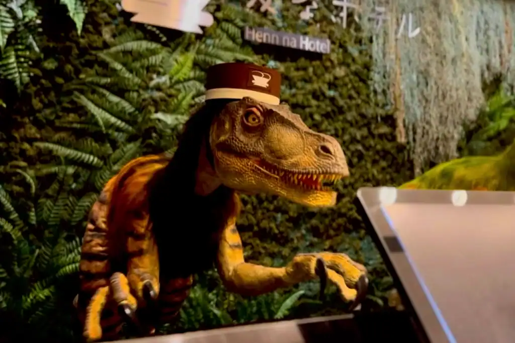 A tan and red animatronic dinosaur head greets guests at Henn-na Hotel, with lobby greenery and visible signage behind.