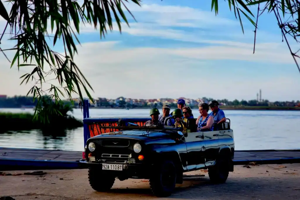 A blue open-top Vietnam War jeep with travellers riding along the riverside near Hoi An, with palm leaves framing the view.