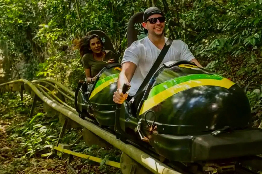 Two riders in a green-and-yellow bobsled wear helmets as they travel through lush Jamaican rainforest at Mystic Mountain.