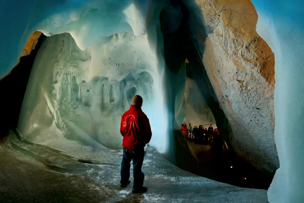 A lone visitor stands in the frozen ice tunnel of Eisriesenwelt, with icy walls and blue light evoking the Hell gateway legends.