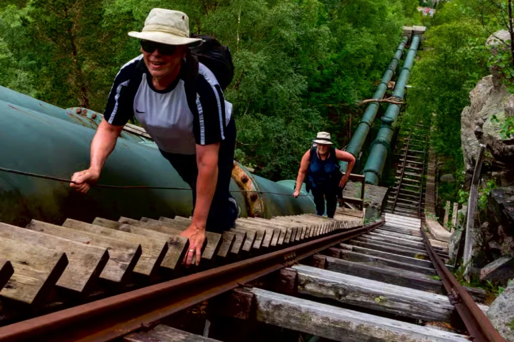 Two hikers climb the Flørli Stairs in Norway, navigating a near-vertical wooden staircase beside green industrial pipes on a 4,444-step route uphill.