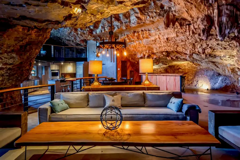 Warmly lit living room inside Beckham Creek Cave Lodge, with a beige sectional sofa, wood coffee table, and rugged stone walls in Arkansas.