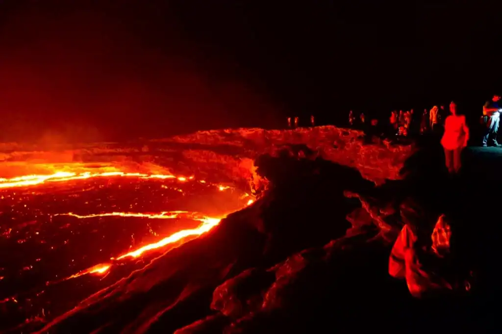 A glowing red lava flow cuts through a dark rocky landscape as onlookers stand along a ridge, facing the Danakil Depression’s hazards.